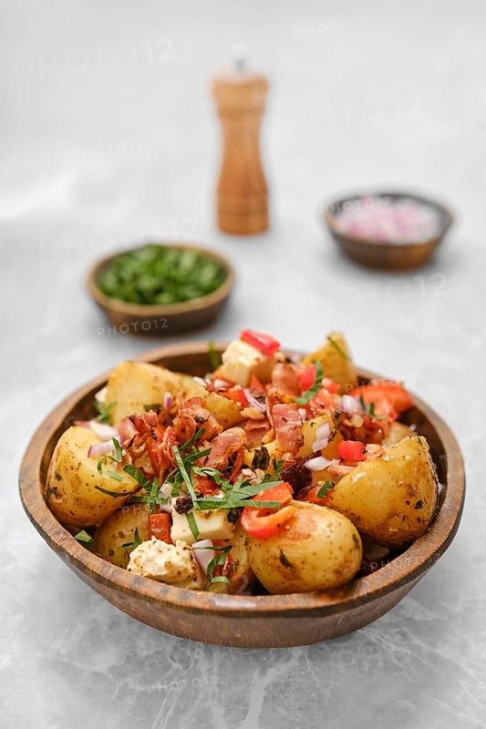 A delicious bowl of roasted new potatoes is garnished with bacon, fresh herbs, chopped onions, and colorful vegetables, creating a vibrant dish perfect for sharing at a family gathering or picnic