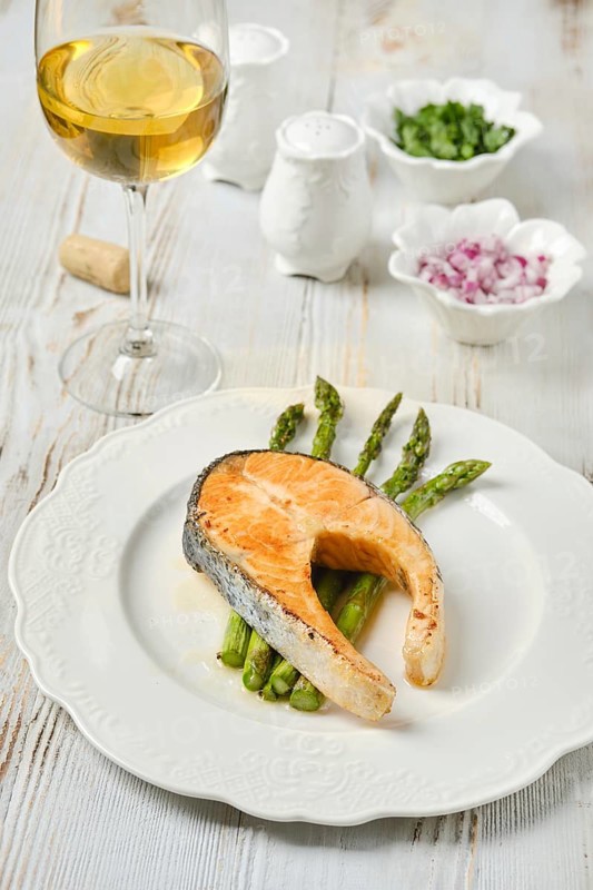 A succulent piece of salmon on a plate with crispy asparagus spears. Accompanying the dish is a glass of white wine. Small bowls of seasonings complete the table setting
