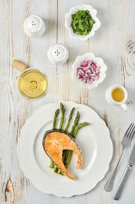 Grilled salmon and tender asparagus on a plate served for dinner. Bowls of chopped onion and herbs accompany a glass of white wine