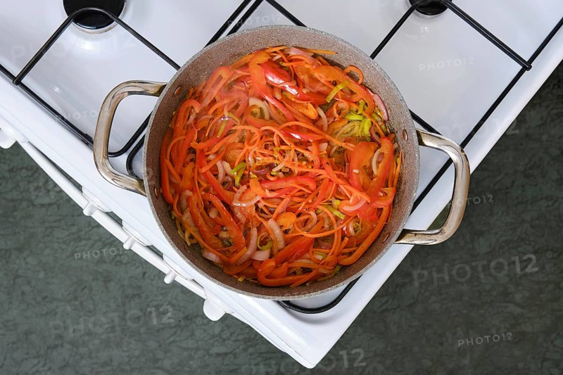 Freshly sliced bell peppers, carrot and onions simmer in a pot on the stove in a cozy kitchen