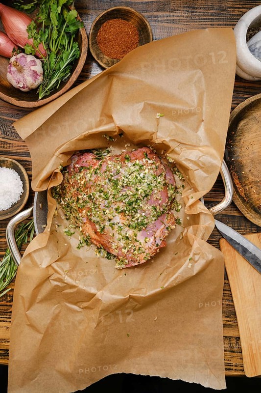 A seasoned turkey thigh in a pan lined with parchment paper, surrounded by fresh herbs, spices, and other ingredients. This cozy kitchen radiates warmth and culinary inspiration