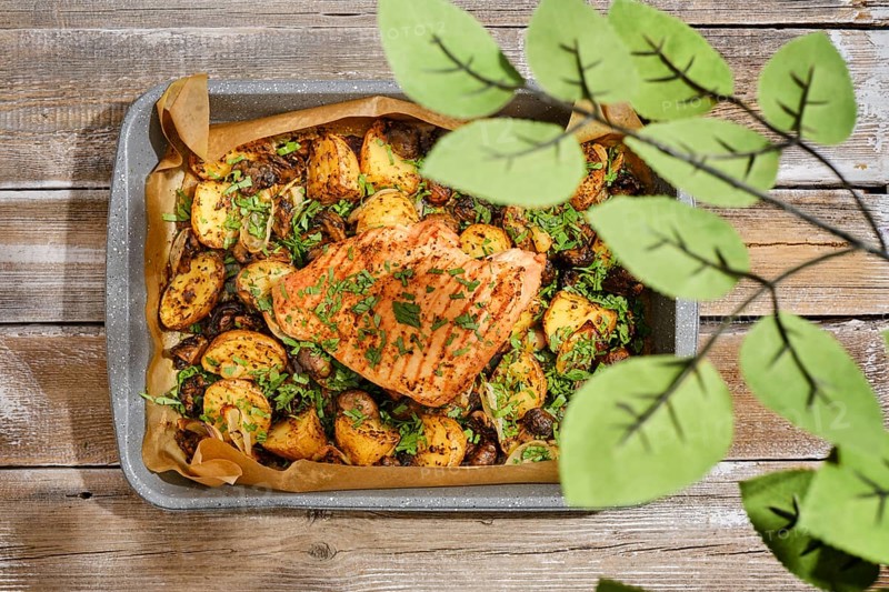 A roasted turkey breast is presented in a baking tray alongside golden potatoes and herbs. The setting features wooden surfaces and green plant leaves, creating a cozy, homely atmosphere