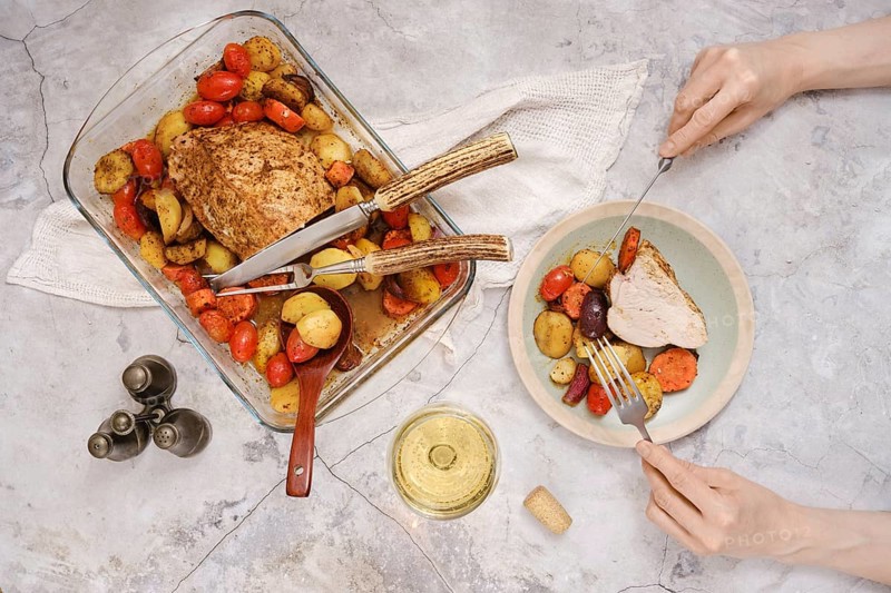 Tender roasted turkey breast rests in a baking dish surrounded by vibrant roasted vegetables. A person prepares to eat a slice alongside assorted veggies, embodying a warm, inviting meal