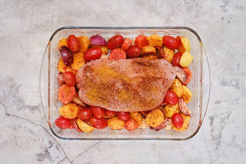 A perfectly seasoned turkey breast rests on a bed of vibrant potatoes, tomatoes and onion ready to be roasted