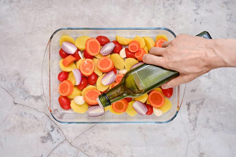 Pouring olive oil on slices of fresh vegetables including tomatoes, carrots, potatoes and onions ready for roasting