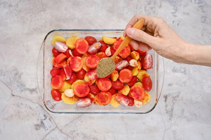 A hand sprinkles oregano over a colorful mixture of tomatoes and other vegetables in a glass baking dish. This preparation is a step for baking turkey breast for a flavorful meal