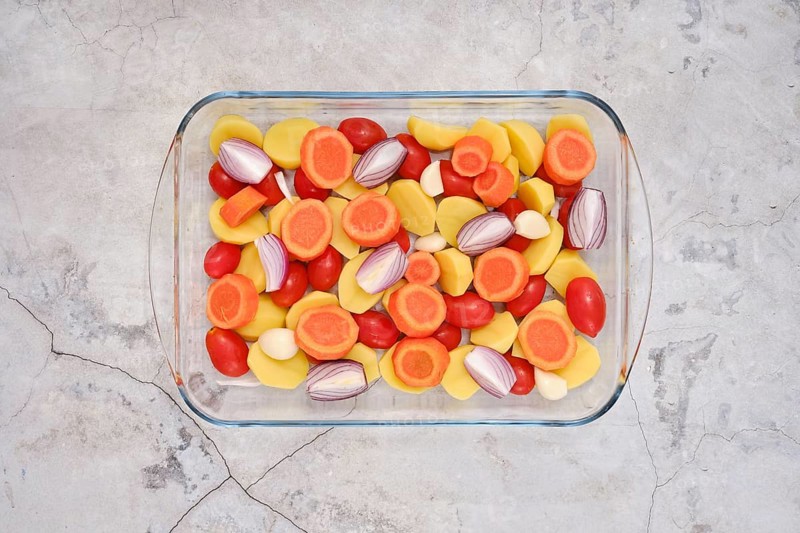 Fresh colourful vegetables like carrots, tomatoes, potatoes, onion and garlic cloves all neatly arranged in a glass baking dish