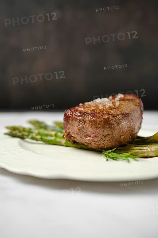 A perfectly cooked filet mignon, seared to golden brown, sits on a decorative plate alongside tender asparagus. The dish is presented in an elegant setting, emphasizing gourmet dining experience