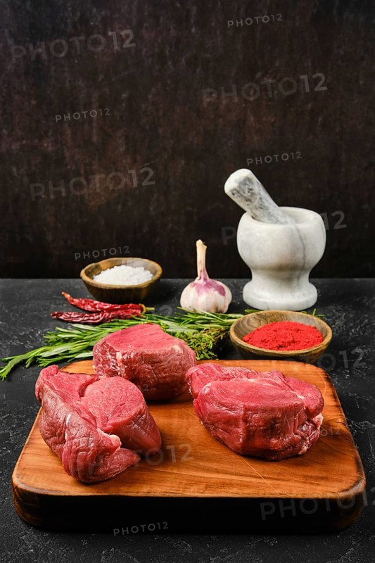 Several cuts of fresh beef tenderloin are arranged on a wooden cutting board, surrounded by spices like paprika powder and salt. Chopped herbs and garlic add color to the kitchen setup, ready for meal preparation