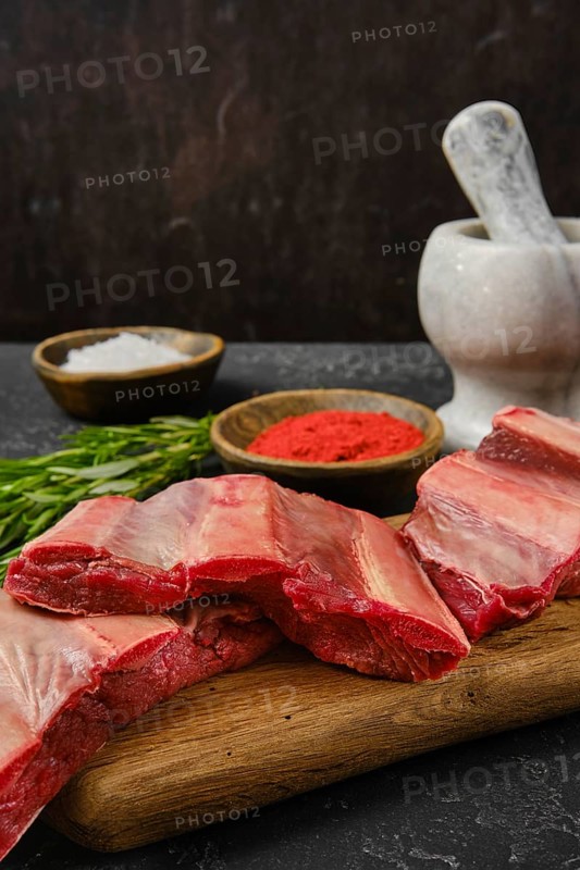 A selection of raw beef ribs is arranged on a wooden cutting board alongside bowls of seasoning. Salt and red seasoning are visible, while fresh herbs add a touch of color and flavor