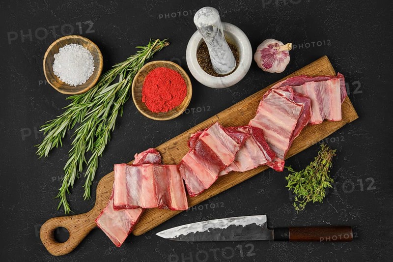 Fresh raw beef short ribs lay on a wooden cutting board alongside bowls of seasoning, herbs, and a knife, indicating a cooking preparation in a home kitchen for a flavorful meal