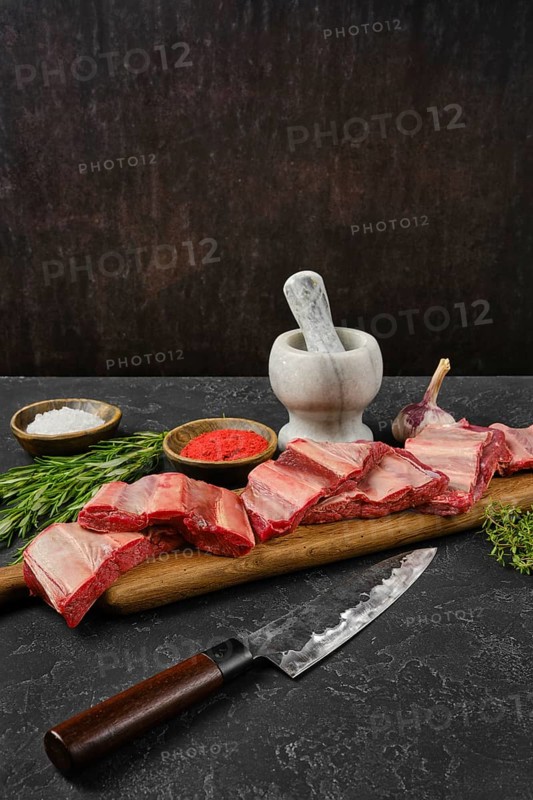 Raw beef ribs are on a wooden cutting board alongside a mortar and pestle. Herbs and spices are arranged nearby, creating an inviting atmosphere for cooking in a rustic kitchen