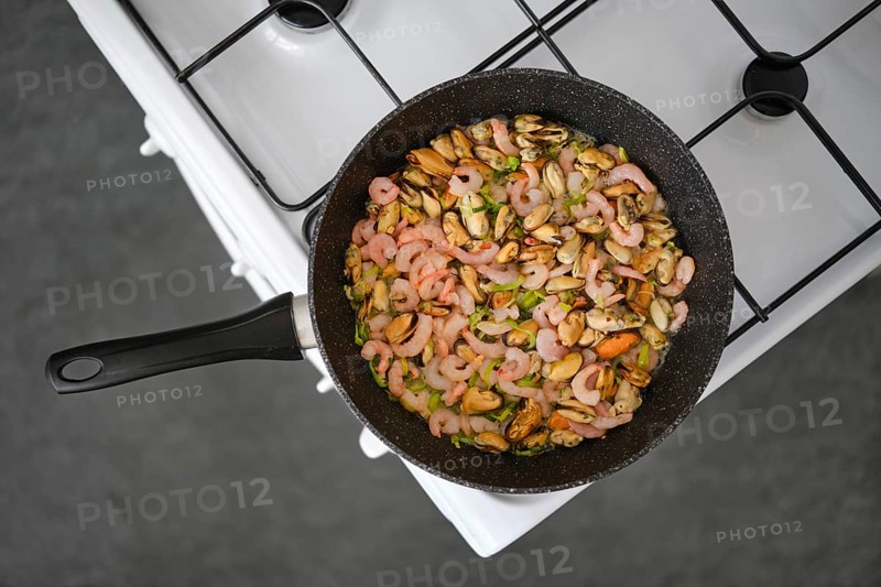 Seafood stir-fry with shrimp and mussels in a black pan, mixed with colourful herbs and spices on a gas stove, overhead view