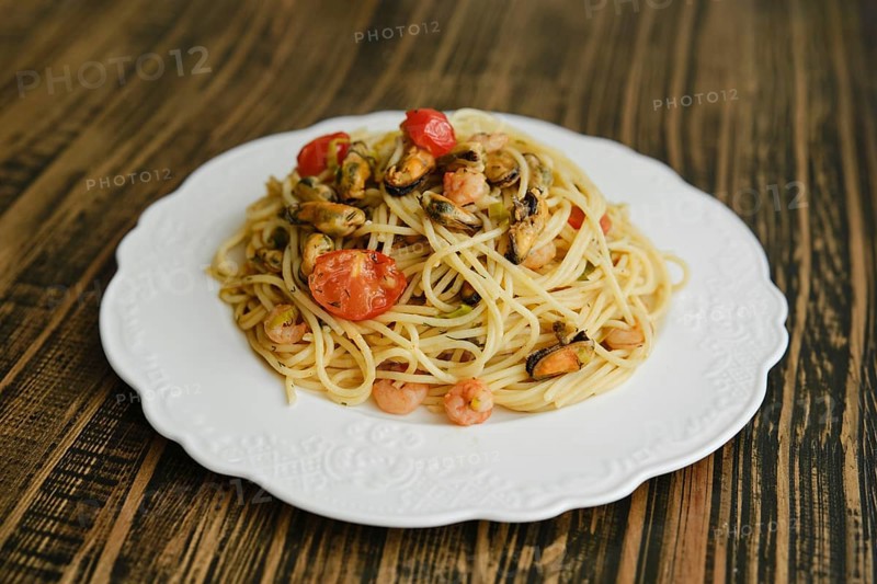 A delicious plate of spaghetti topped with shrimp and mussels, complemented by fresh cherry tomatoes and herbs, sits on a beautifully designed white plate against a rustic wooden table background