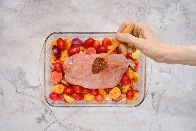 A hand sprinkles a blend of spices over a raw turkey breast resting on a bed of vibrant red and yellow potatoes in a glass baking dish, ready for a flavorful cooking process