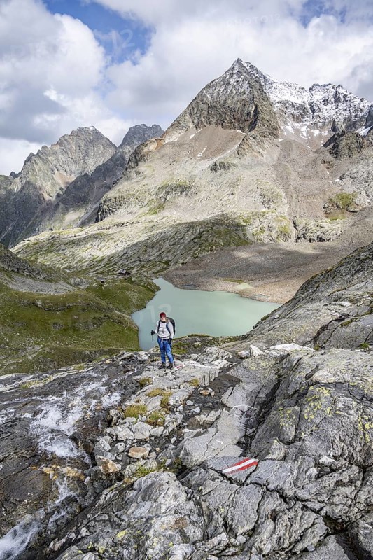 Mountaineer on a rocky hiking trail, view of the turquoise-blue mountain lake Großer Gradensee, behind her the summit of the Petzeck, Hoher Perschitzkopf and Kruckelkopf, Schober Group, Hohe Tauern National Park, Carinthia, Austria
