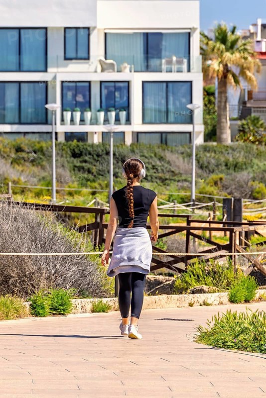 Young woman walking outdoors on a footpath in a residential area and listening to music with headphones from behind, Mallorca, Spain