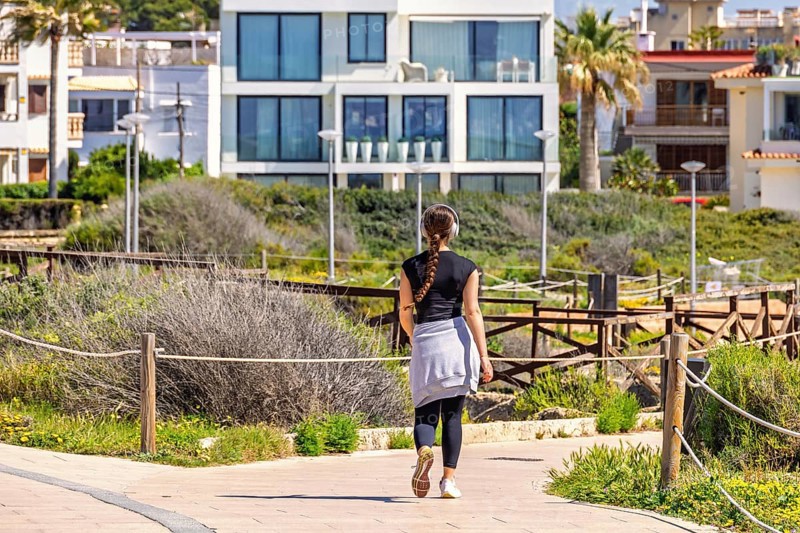 Young woman walking outdoors on a path in a residential area and listening to music with headphones from behind, Mallorca, Spain