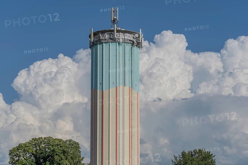 A tall water tower, painted with blue and white stripes, sits enthroned in a green landscape. Fluffy white clouds fill the bright blue sky, creating a picturesque scene during daylight hours. Bas rhin, Alsace, grand est, France