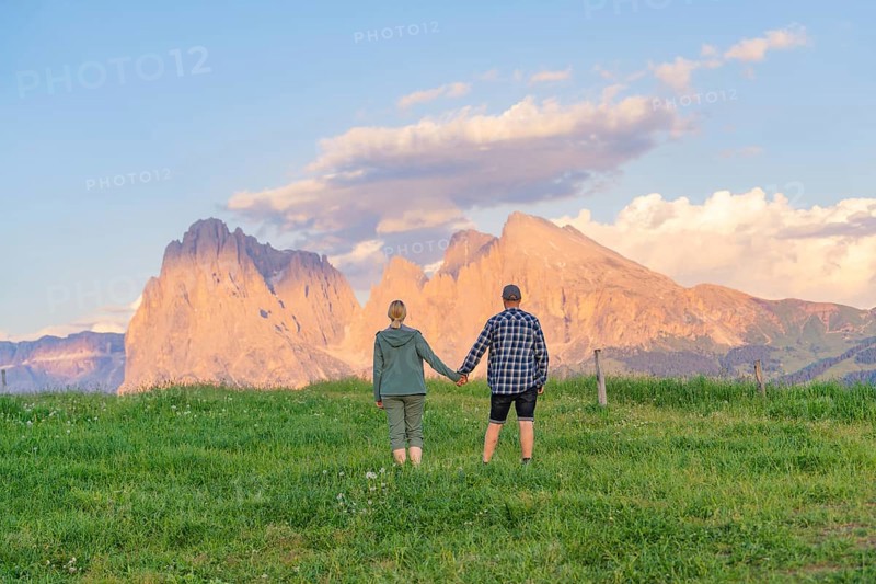 A couple stands hand in hand in a meadow in front of an impressive mountain landscape, Alpe di Siusi, Dolomites, South Tyrol, Italy