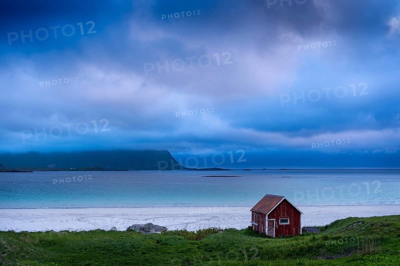 A red wooden hut on the sandy beach of Ramberg (Rambergstranda), sea and mountains. At night at the time of the midnight sun. Clouds in the sky. Early summer. Ramberg, Flakstadoya, Lofoten, Norway