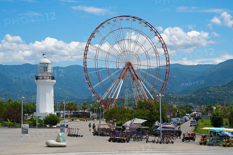 Big Ferris wheel and white lighthouse with mountains in the background under blue sky, amusement park, Batumi, Black Sea, Ajara region, Adjara, autonomous republic, Georgia