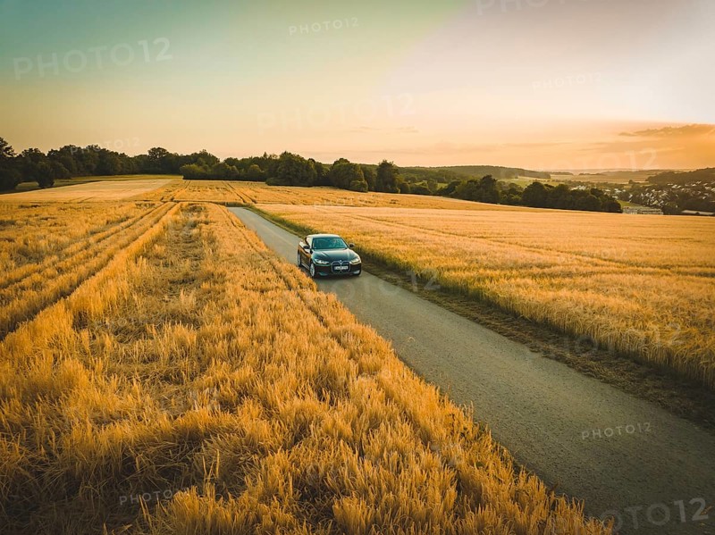 A car drives on a country road between golden fields at sunset, BMW i7, Deer E-Carsharing, Calw, Germany