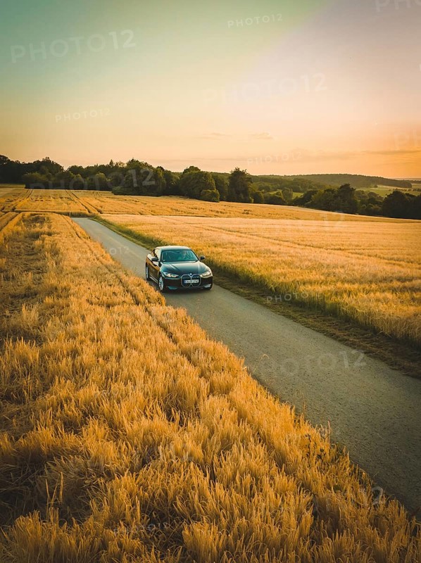A car drives along a road surrounded by vast fields of corn with a colorful sunset in the background, BMW i7 electric car, deer e-car sharing, Calw, Germany