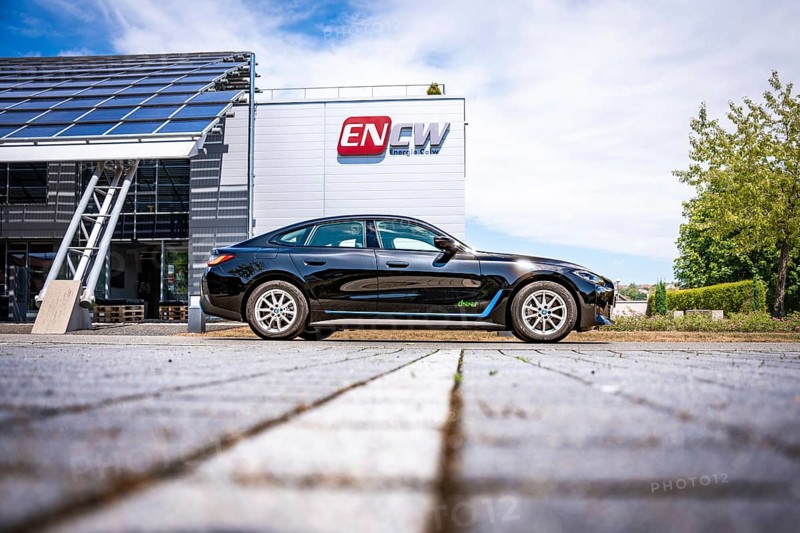 Black car on the side of a modern building with ENCW logo on a paved path, BMW i7 electric car, Deer e-Carsharing, Calw, Germany