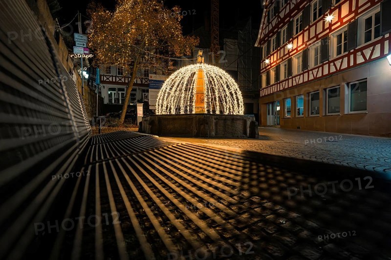 Night view of a festively illuminated fountain with shadows cast in a historic street, Calw, Black Forest, Germany