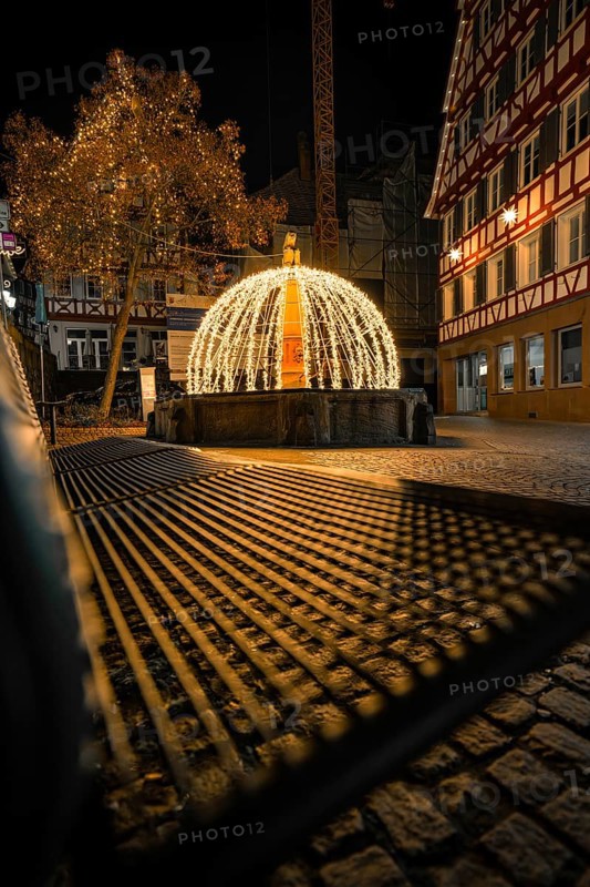 Festively illuminated fountain with lights and shadows on a paved road at night, Calw, Black Forest, Germany