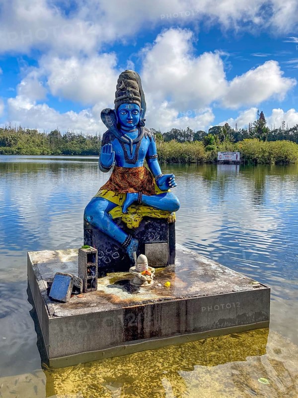 Statue sculpture of deity Shiva on the shore of stands in sacred Hindu lake Ganga Talao at religious site largest Hindu sanctuary for religion Hinduism outside India for believing Hindu Hindus, in the background Sacred Lake Ganga Talao crater lake in crater of extinct volcano, Grand Bassin, Le Pétrin, Mauritius