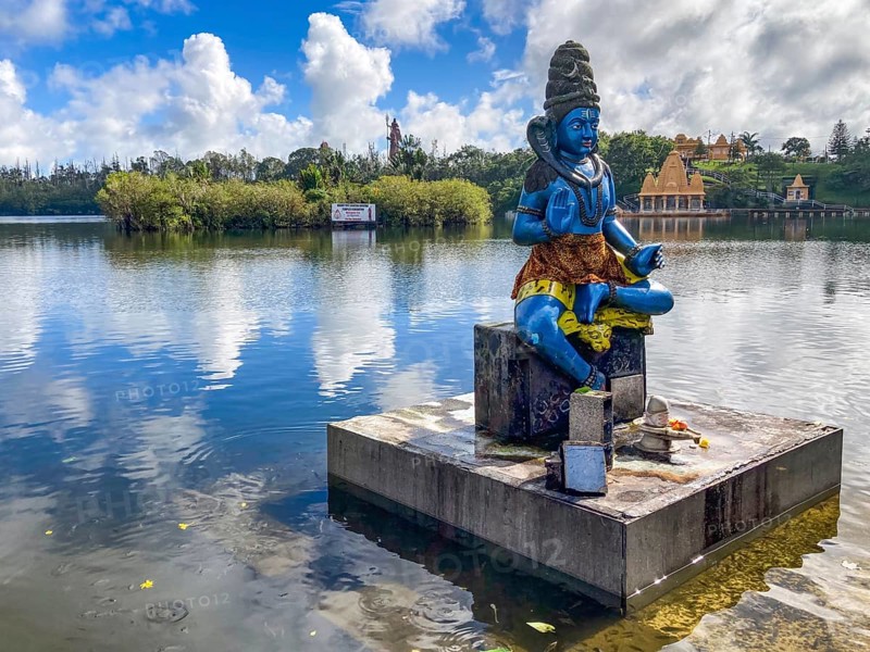 In the foreground statue sculpture of the deity Shiva stands in sacred Hindu lake Ganga Talao at religious site largest Hindu sanctuary for religion Hinduism outside India for believing Hindu Hindus, in the background Sacred Lake Ganga Talao crater lake in crater of extinct volcano, Grand Bassin, Le Pétrin, Mauritius