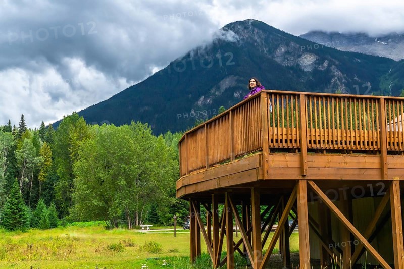 Tourist standing on a wooden deck, admiring the breathtaking view of mount robson, the highest peak in the canadian rockies, surrounded by lush green forest and clouds