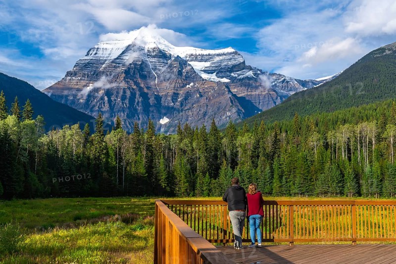 Two tourists standing on a wooden deck are enjoying the breathtaking view of mount robson, the highest peak in the canadian rockies, surrounded by lush green forest and a vibrant meadow