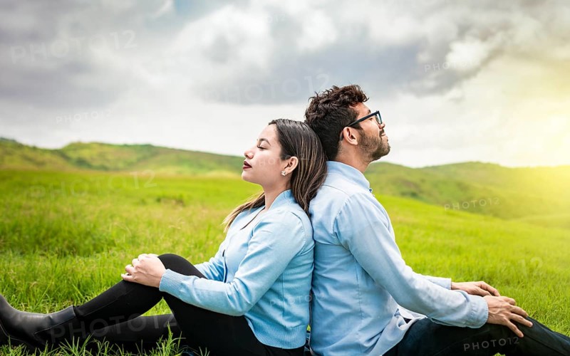 A couple sitting with their backs to each other on the grass, Wedding couple in the field sitting with their backs to each other looking towards the camera