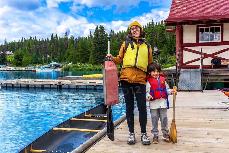Mother and son holding paddles and wearing life jackets, standing on a wooden dock beside a canoe at maligne lake in jasper national park, alberta, canada