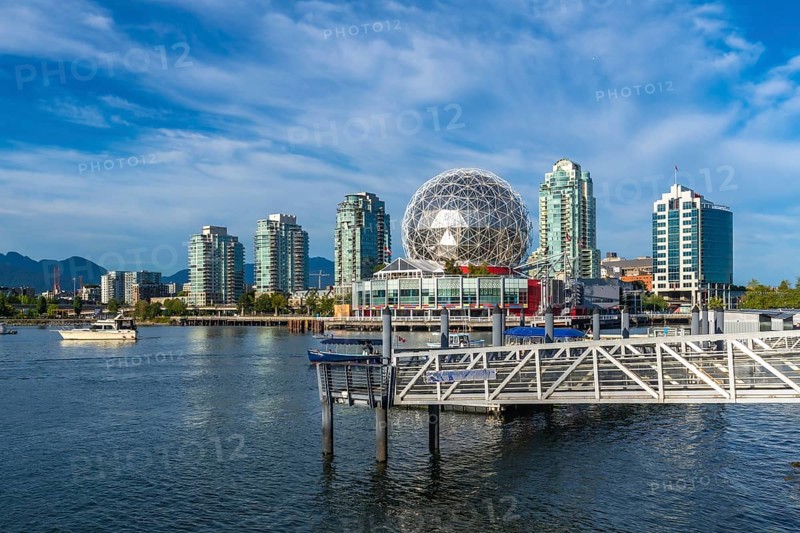 Modern buildings and science world are reflecting in the calm waters of false creek, vancouver, with mountains in the background under a beautiful blue sky on a sunny day