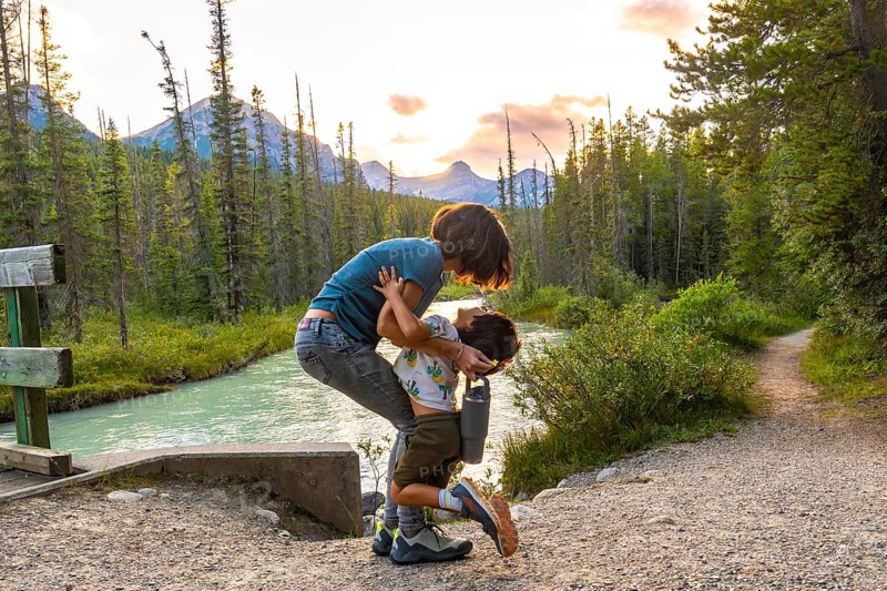 Mother embracing her son by the riverside in banff national park, surrounded by the stunning canadian rockies at sunset, capturing a heartwarming family moment in nature