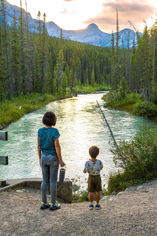 Mother and her son are enjoying the breathtaking view of a pristine river flowing through a lush forest, surrounded by the majestic canadian rockies at sunset in banff national park, alberta