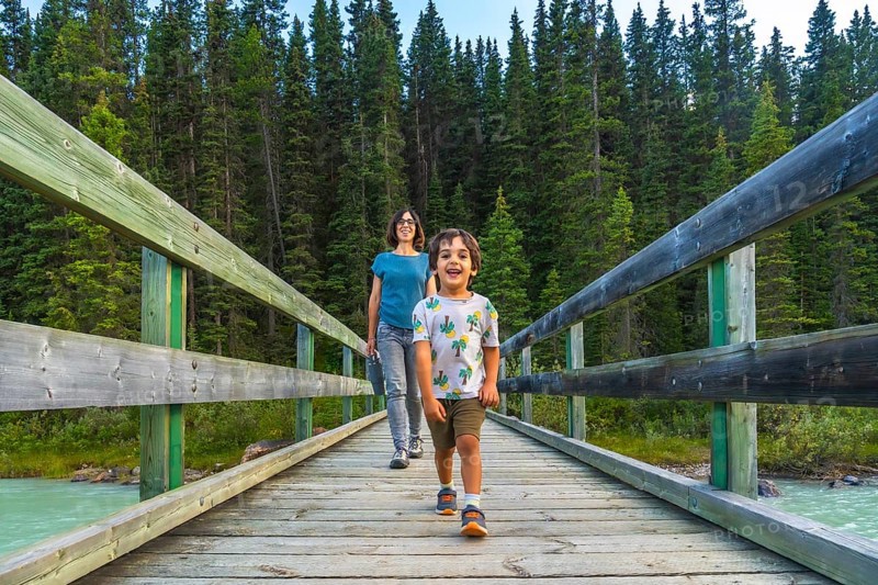 Mother and son enjoying a leisurely walk across a wooden bridge, surrounded by the stunning scenery of pine trees and turquoise river in banff national park, canadian rockies