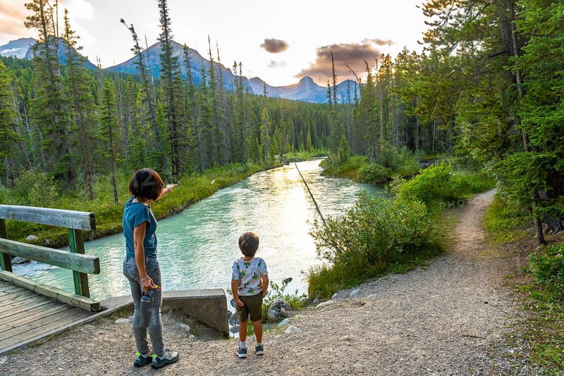Woman points towards the majestic mountains in the distance as she and her son stand near a tranquil river in banff national park, enjoying the serene sunset view