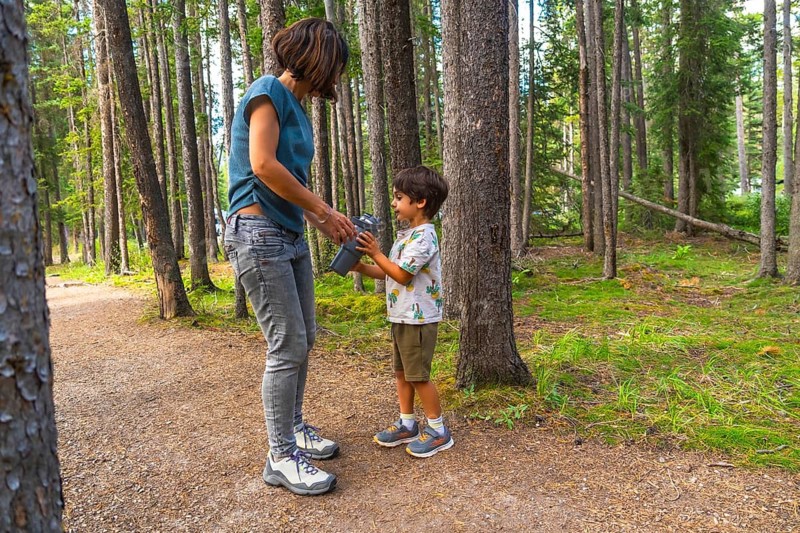 Mother hands a water bottle to her son during a hike on a sunny day surrounded by the lush green trees of banff national park in the canadian rockies