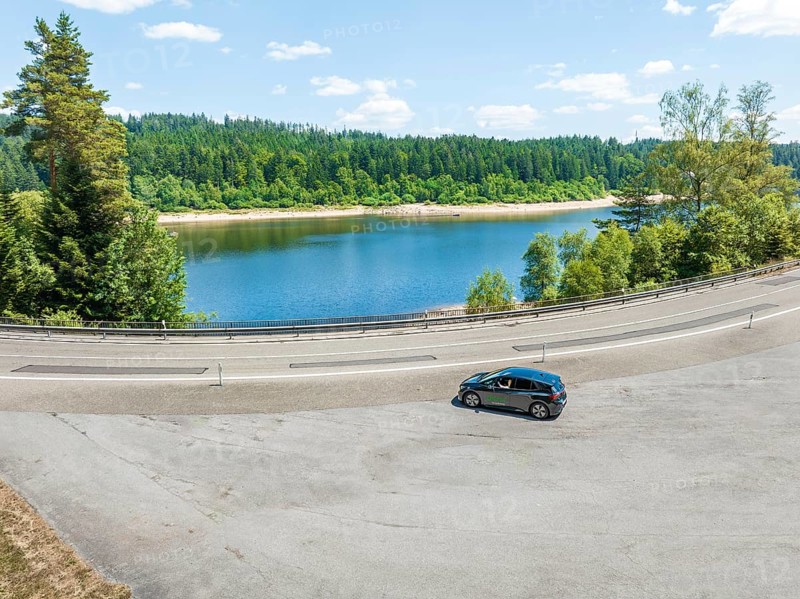 A black car drives along a lake, surrounded by a forest, in clear skies, deer e-car sharing, Cupra electric car, Black Forest, Germany
