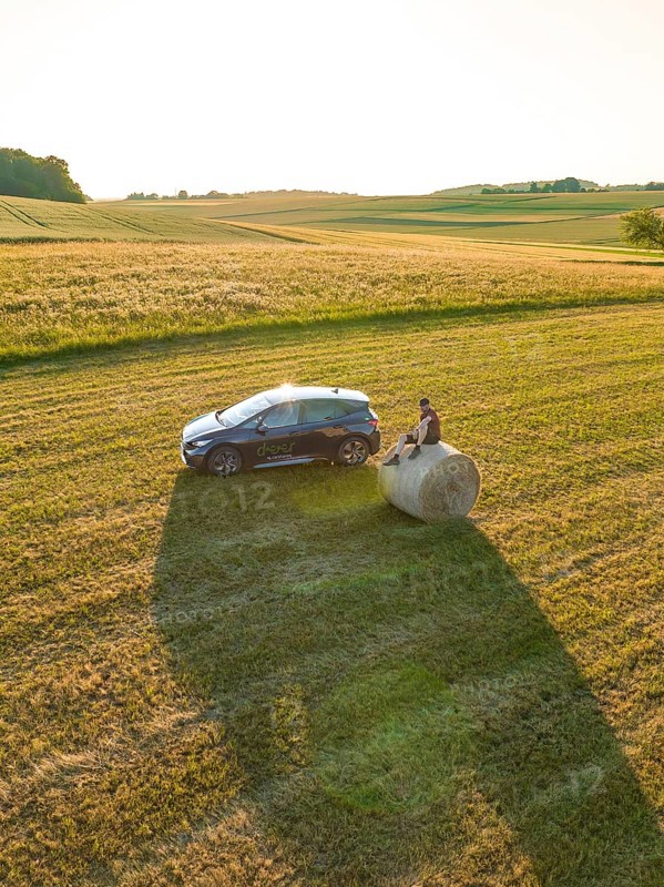 Car with person on a hay bale in a wide field at sunset, deer e-car sharing, Cupra electric car, Gechingen, Germany