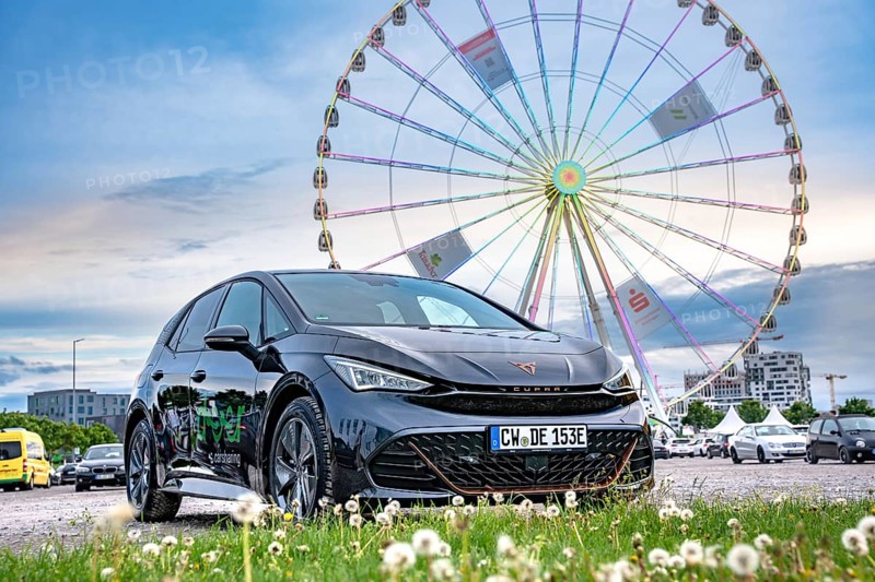 Black car in front of a Ferris wheel in a field with blue sky and clouds, Deer E-Car Sharing, Cupra electric car, Böblingen, Germany