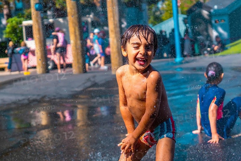 Cheerful child with wet hair and colorful swimming trunks laughing and playing in a city park splash pad, enjoying refreshing water sprays on a sunny day