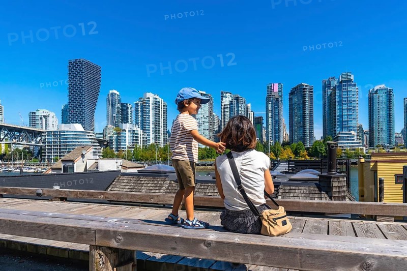 Sitting on a wooden bench at granville island market, a mother and son admire the stunning vancouver skyline under a clear blue sky on a sunny day