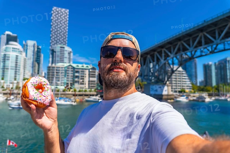 Tourist taking a selfie while holding a donut with burrard bridge and the cityscape of vancouver in the background, during a sunny summer day