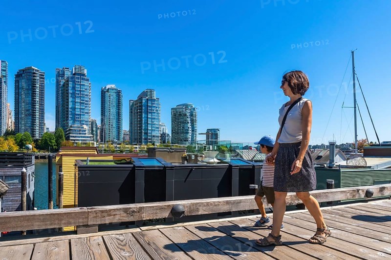 Mother and her son enjoying a sunny summer day walking along the wooden boardwalk of granville island market with the cityscape of vancouver in the background